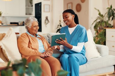 A senior woman smiling while sitting on a couch with a caregiver in scrubs, reviewing a tablet together in a cozy home setting.