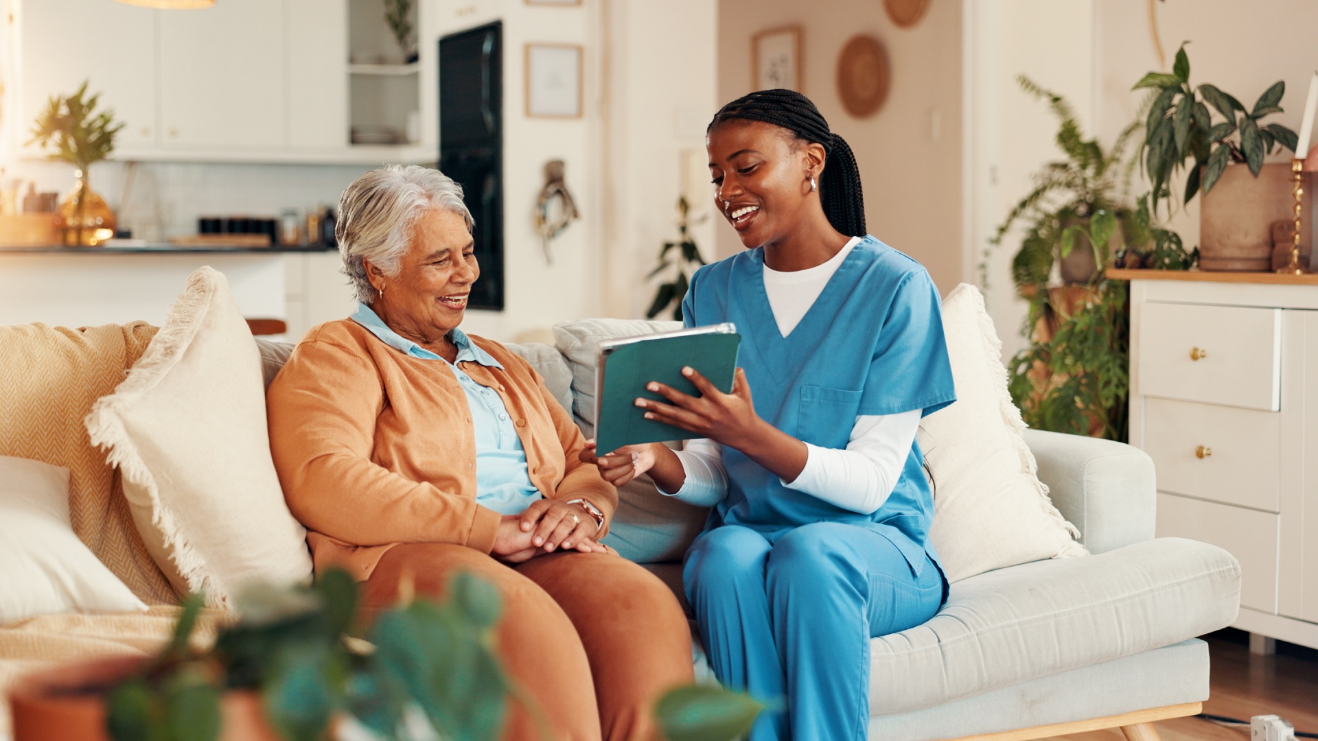 A senior woman smiling while sitting on a couch with a caregiver in scrubs, reviewing a tablet together in a cozy home setting.